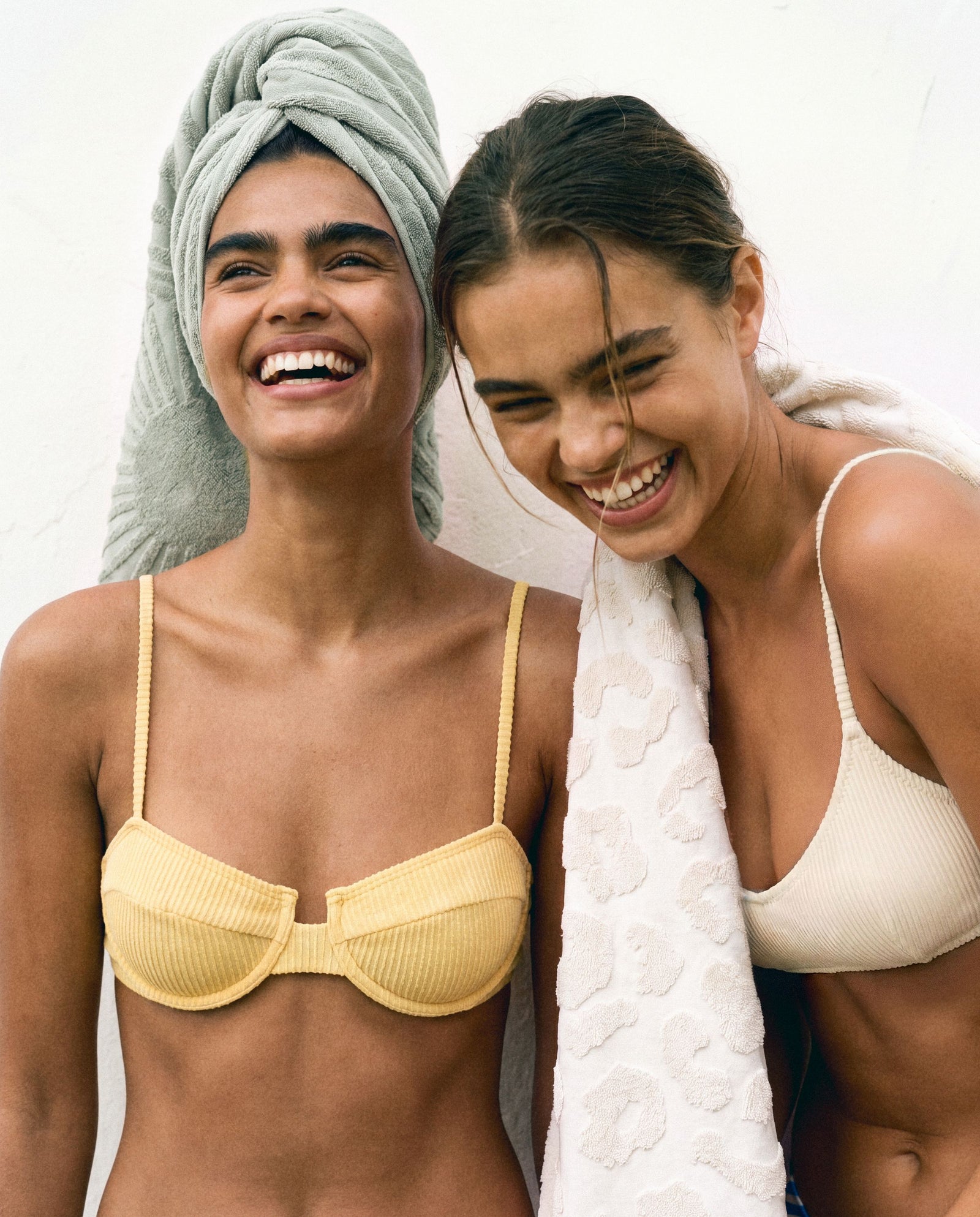 Two friends laughing together, wrapped in Ruby Mint towels against a sunlit white wall