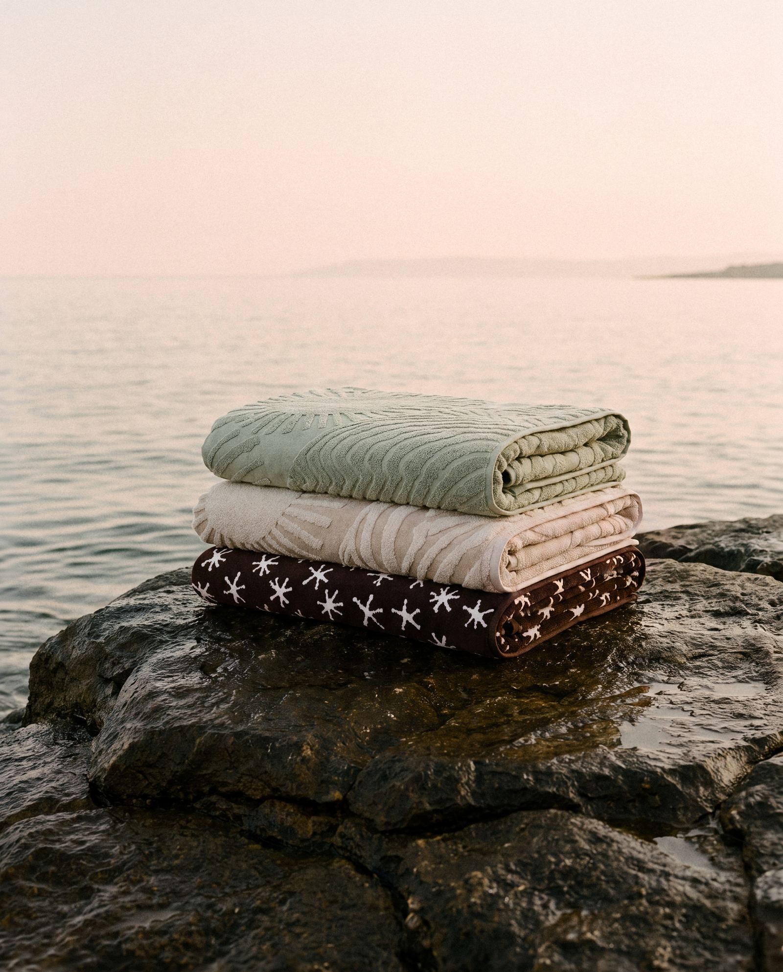 Three folded Ruby Mint towels stacked on a sea rock at dusk